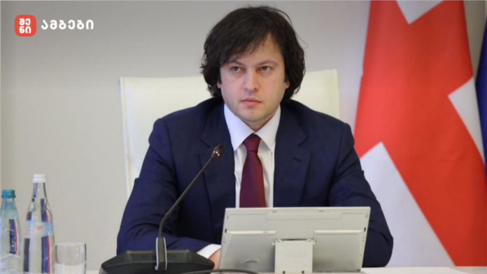 Man in a navy suit and red tie sits at a conference table speaking into a microphone, with a tablet in front and a Georgian flag in the background.