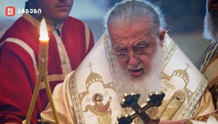 Eastern Orthodox priest wearing ornate vestments and holding a cross during a candlelit ceremony with attendants nearby, blue-toned background.