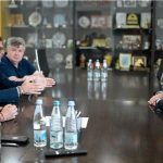 შეხვედრაზე „Develey“-სა და „მარნეულის სასურსათო ქარხნის“ თანამშრომლობის ფარგლებში, „მარნეულის სასურსათო ქარხანაში“ ახალი საწარმოო სივრცის მოწყობის მიმდინარეობა და სამომავლო გეგმები განიხილეს Four professionals sit around a dark wooden conference table in a meeting, with water bottles and a smartphone on the table; shelves with awards in the background.